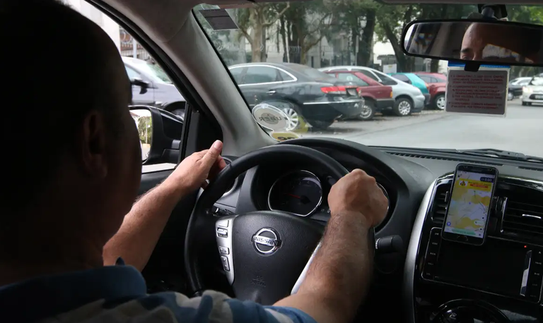 Motorista dirige por avenida olhando aplicativo de corrida. Foto: Rovena Rosa/Agência Brasil